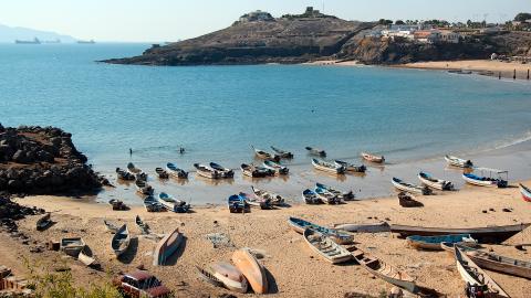 Photo of azure blue water and a beach with rowboats sitting in the sand and in the shallow water. The rocky coastline wraps around with a village up on cliffs in the distance.