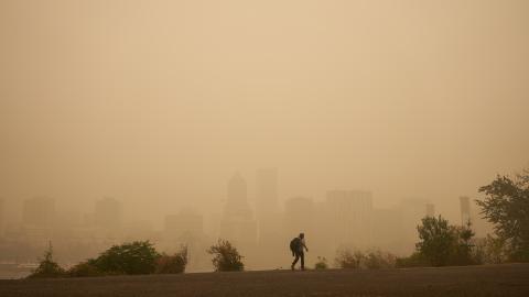 Smoky skies turn the Portland skyline hazy and orange-hued. A person with a backpack walks in the foreground of the image.