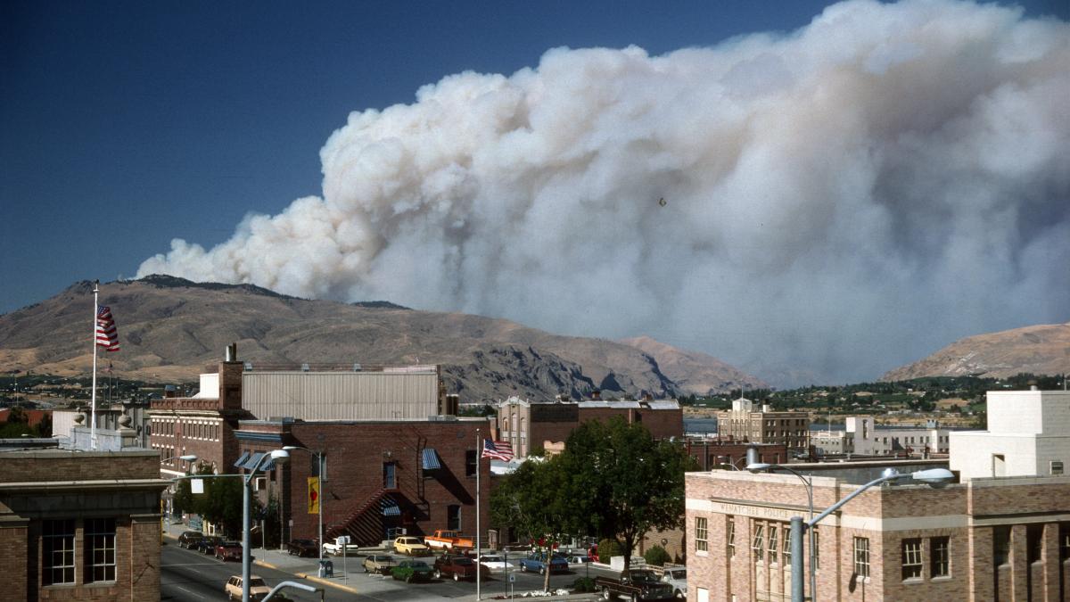 A plume of wildfire smoke behind the center of a small town showing brick buildings and cars.