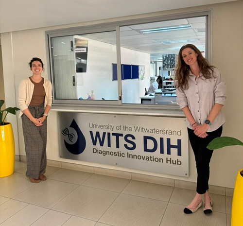 Alaina Olson and Rachel Wood stand on either side of an internal window looking into a lab with a sign below reading "University of the Witwatersrand Diagnostic Innovation Hub - WITS DIH"