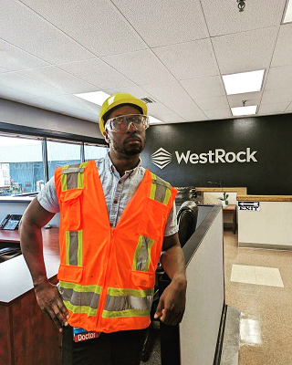 A man in safety vest, hardhat and safety glasses stands indoors with a sign behind him reading WestRock.