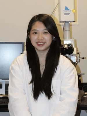 Cayen Rong stands smiling in front of a computer monitor and a microscope.