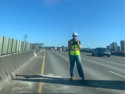 Chamonix Reynolds stands on a highway bridge with cars in the background. She is holding a camera and wearing a helmet and safety vest.