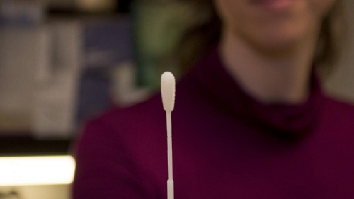 An oral swab held up by a person who is in the blurry background, with only chin, neck, shoulders and chest visible.