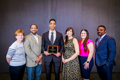 Six people stand together smiling with a stage curtain behind them. Elijah Scott is third from left holding an award plaque.