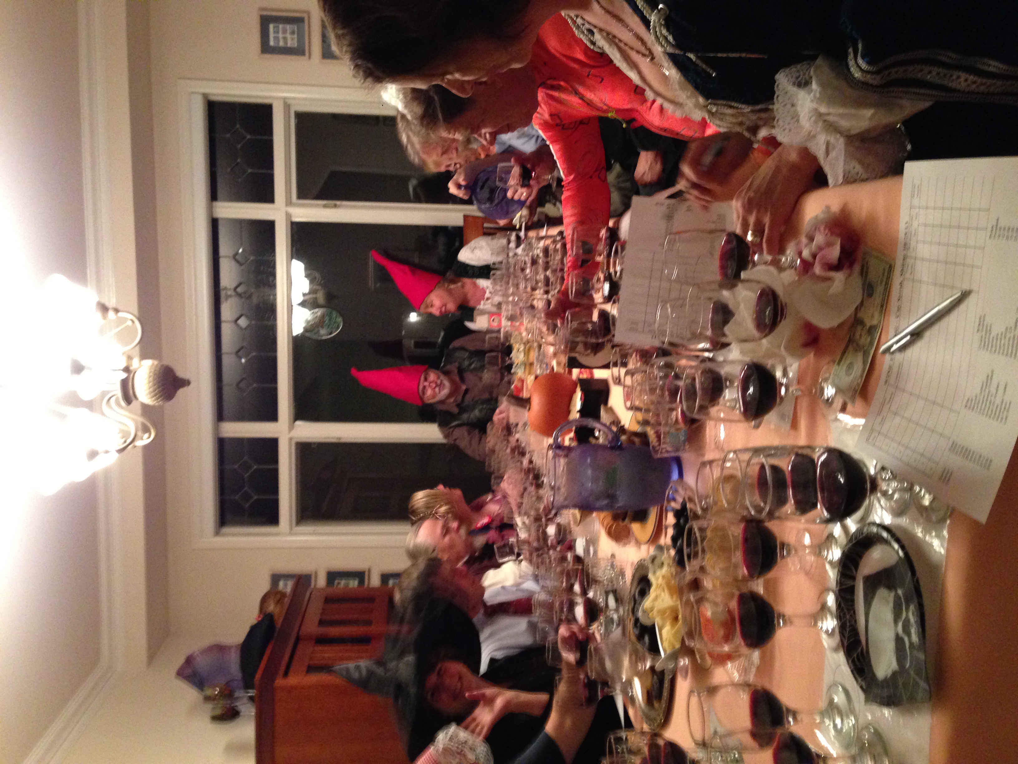 A group of 10 people sit around a dining table covered with many wine glasses with small amounts of wine in them, a pitched of water, a pumpkin and other foods. One person is wearing a black witch hat and two people are wearing red gnome hats.