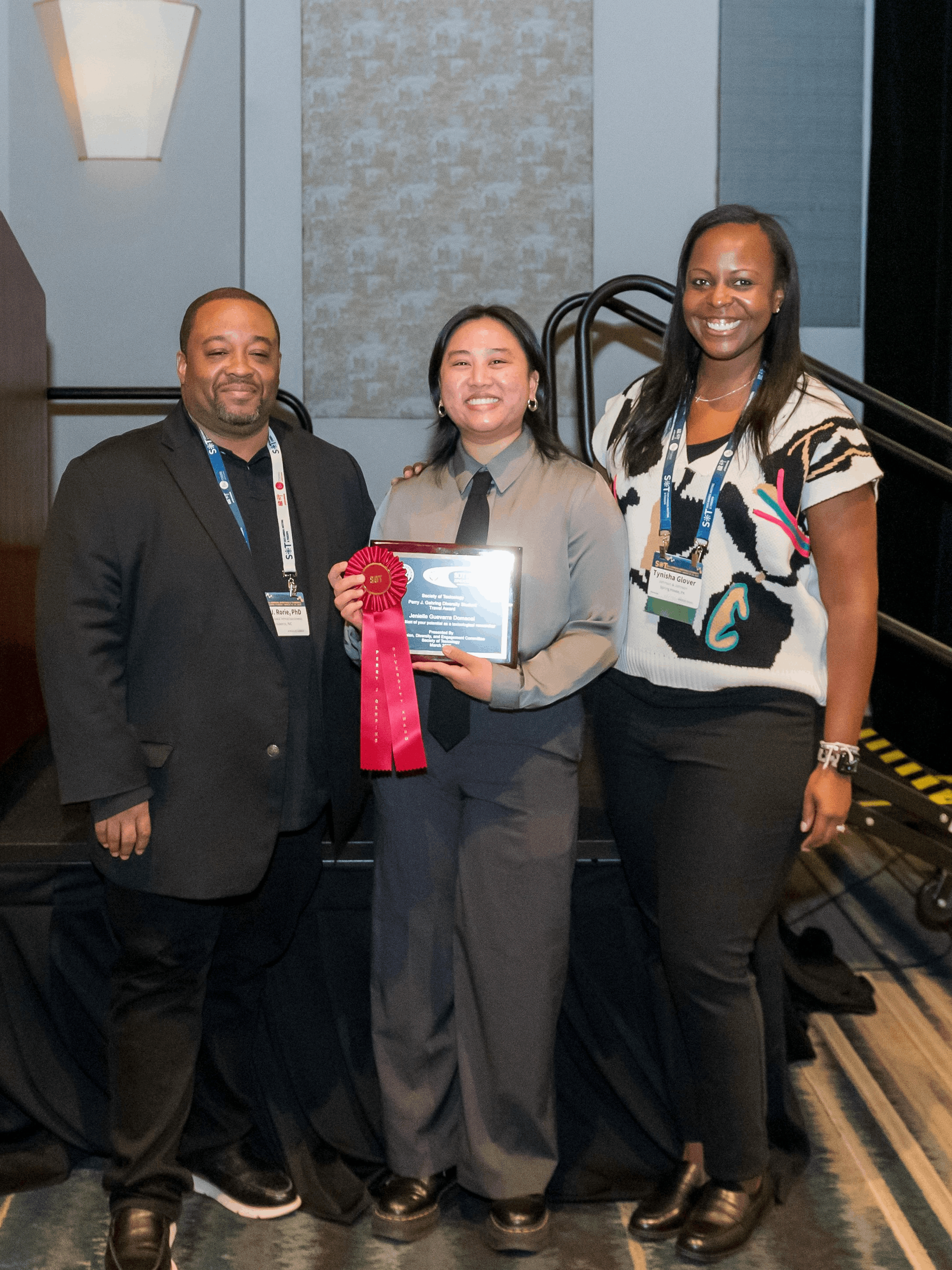 Jenielle Domaoal stands in between two people while holding a plaque and red ribbon for her award.