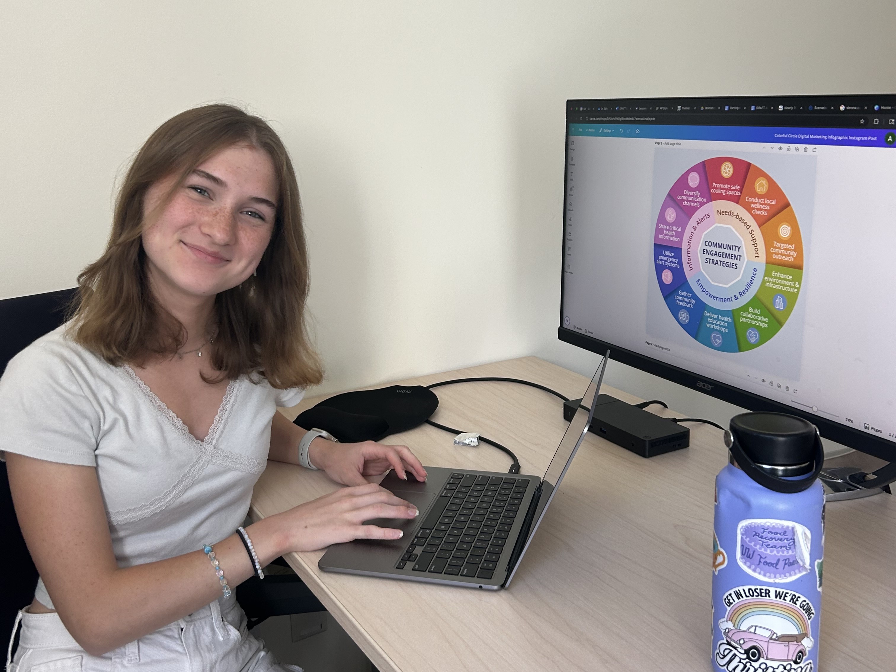 Woman with light brown hair sits at desk with computer and water bottle.