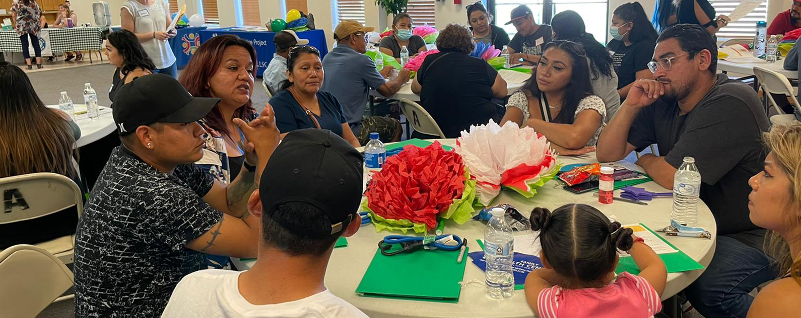 A table of seven farmworkers and a child sit in the foreground having a discussion, with other tables of people in the background.