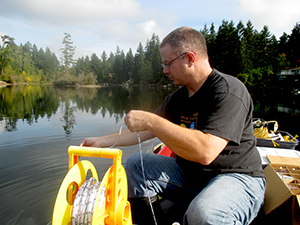 UW research technologist Corey King measures the water transparency of Lake Killarney. He sits in a boat with the lake and trees int he background, working with a large spool of rope. Photo: James Gawel.