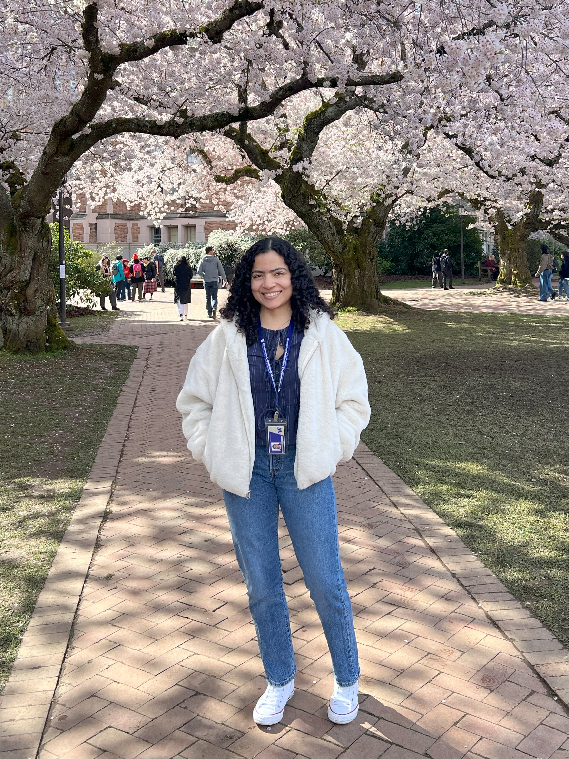 Student with long black curly hair wears a white jacket and stands among cherry trees.