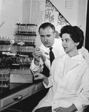 A black and white photo of a man and woman in a lab with glassware. The man is looking down at a test tube in his hands.