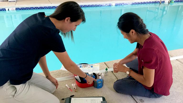 Ali, pictured on the left, assists with a swimming pool inspection. She and another woman sit on the concrete edge of a chlorinated pool with testing equipment and a clipboard.