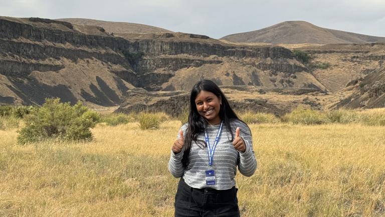 Student with long black hair stands in front of a rural landscape with grasses and steep bluffs in background. She wears an ID badge on a lanyard and holds two thumbs up.