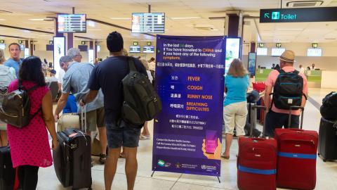 A group of travelers with suitcases stand in line at an airport near a sign about the novel coronavirus