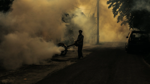 A worker performs anti-mosquito fogging. The worker is in shadow on a dark street with billows of smoke rising up in the air.